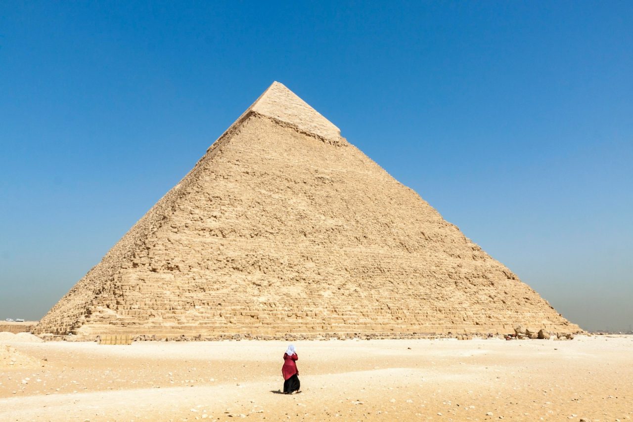 woman in black jacket walking on brown sand near pyramid under blue sky during daytime